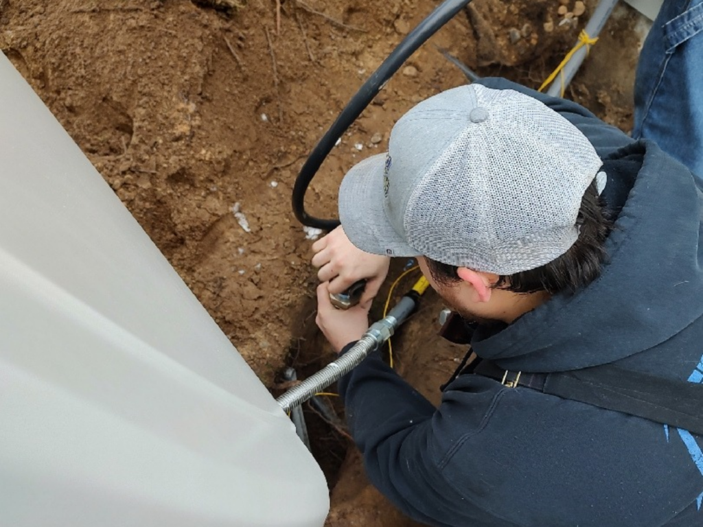 A plumber wearing a gray cap and black hoodie installs outdoor gas line piping for a generator.