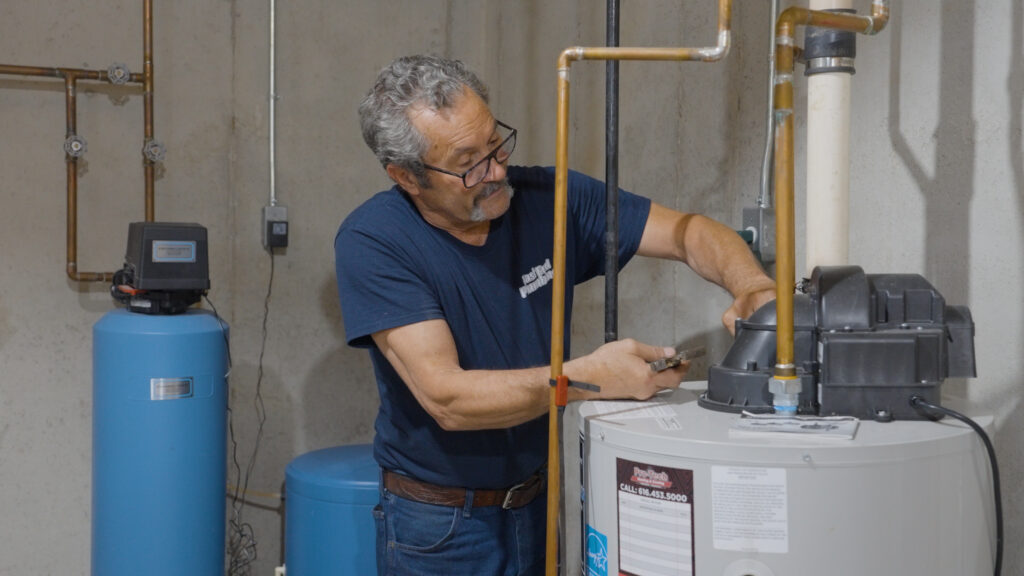 Man wearing glasses uses a wrench to adjust a component on a water heater in a basement utility room with exposed pipes.